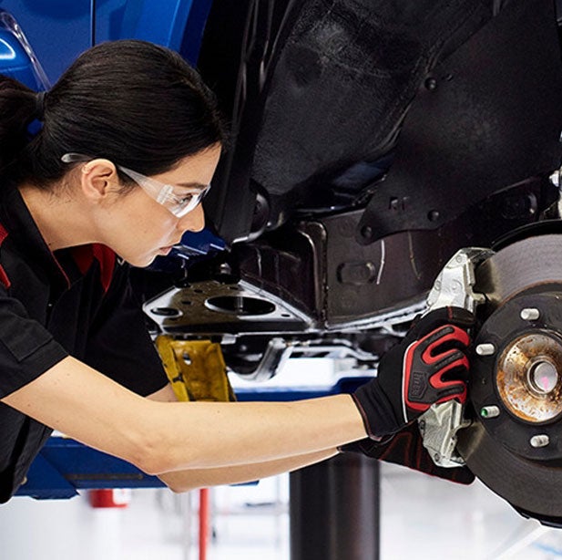 Female mechanic in safety glasses and gloves working on a car's brake disk and caliper assembly.