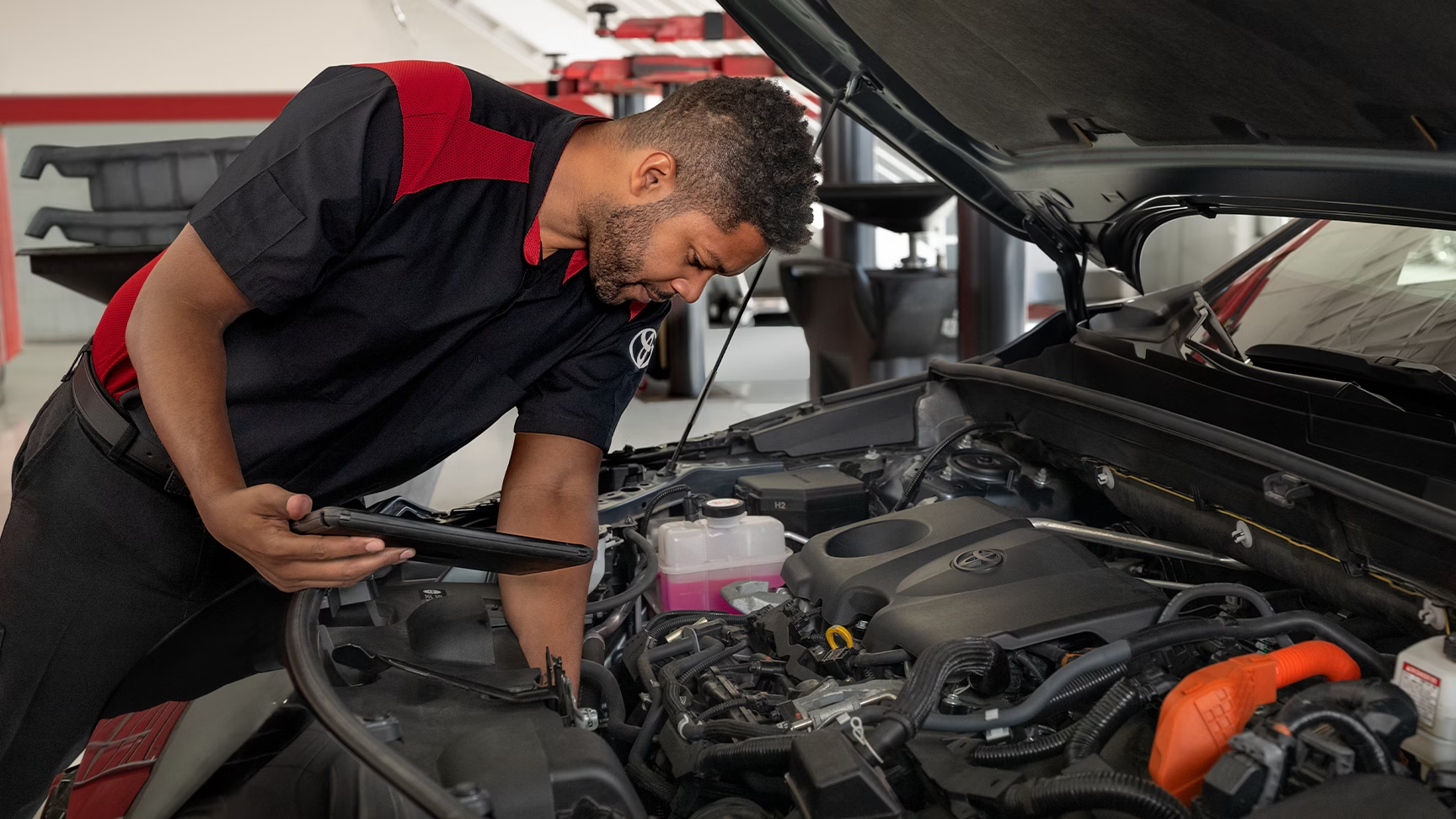 A mechanic technician holding a tablet examines a car engine with the hood open in a dealership service bay.